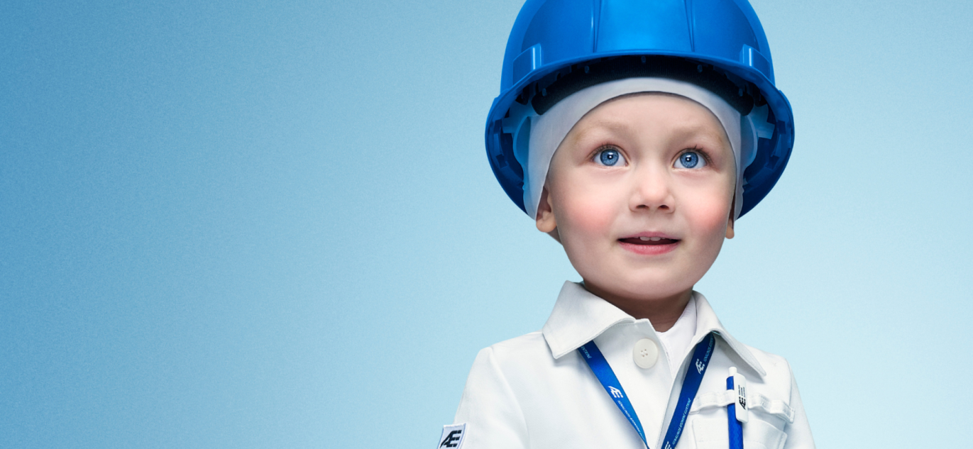 Mažas vaikas vilkintis baltą uniformą ir dėvintis mėlyną apsauginį šalmą su Ignalinos atominės elektrinės logotipu.