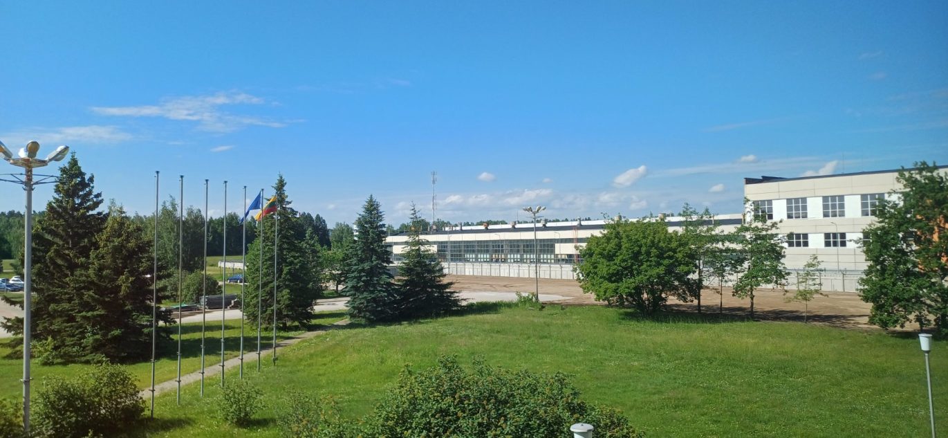 Ignalinos atominės elektrinės teritorija vasaros metu, su matomomis vėliavų stiebais, želdiniais ir administraciniu pastatu fone.