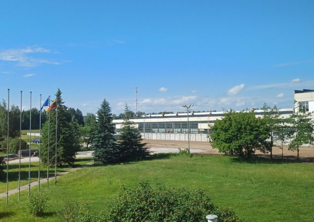 Ignalinos atominės elektrinės teritorija vasaros metu, su matomomis vėliavų stiebais, želdiniais ir administraciniu pastatu fone.