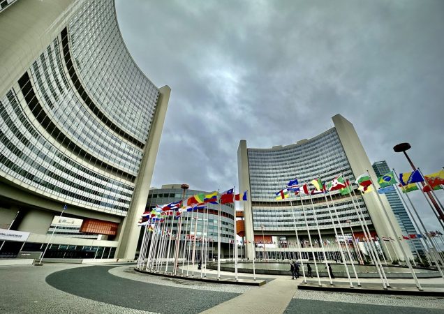 Tarptautinės atominės energijos agentūros (TATENA) būstinė Vienoje, apsupta pasaulio šalių vėliavų, su modernia architektūra ir niūriu dangumi fone.