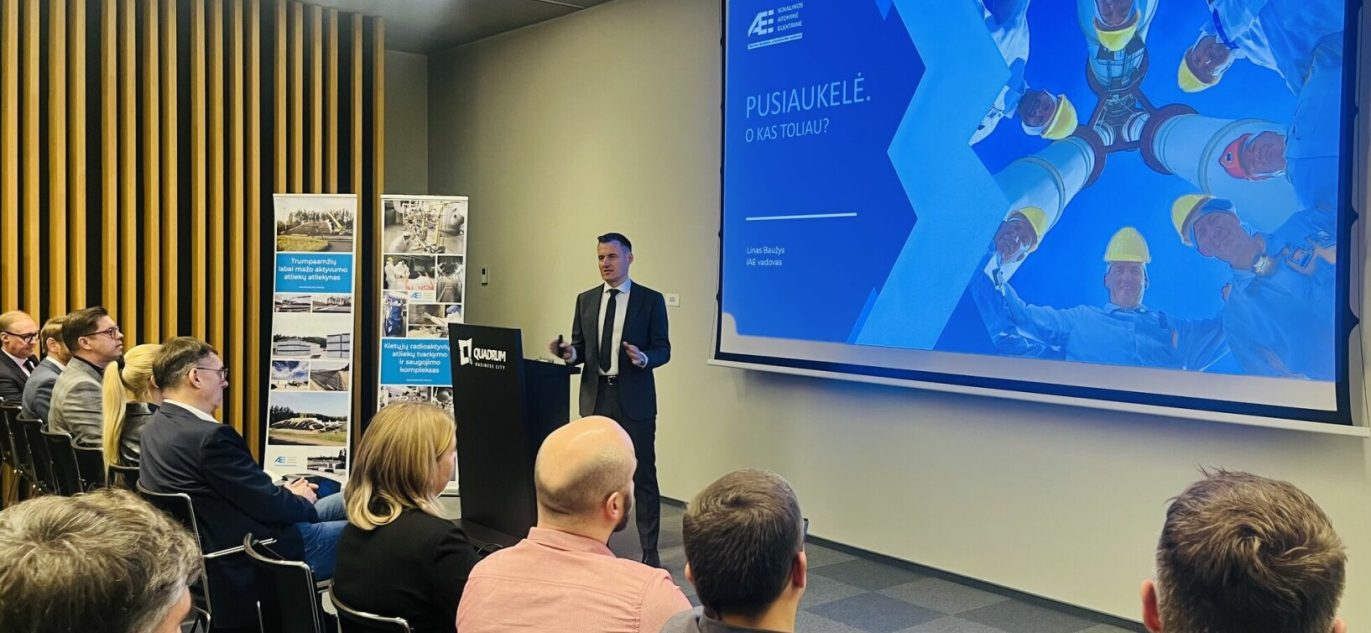Pranešėjas pristato skaidrę tema „Pusiaukelė. O kas toliau?“ konferencijoje, kurioje dalyvauja Ignalinos atominės elektrinės atstovai ir kviestiniai svečiai. Auditorija įdėmiai klauso.