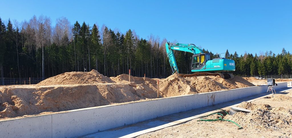Statybvietėje ekskavatorius pila smėlį ant žemės sankasos šalia naujai įrengto betoninio pamato, fone matosi miško juosta ir giedras dangus.