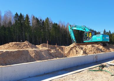 Statybvietėje ekskavatorius pila smėlį ant žemės sankasos šalia naujai įrengto betoninio pamato, fone matosi miško juosta ir giedras dangus.