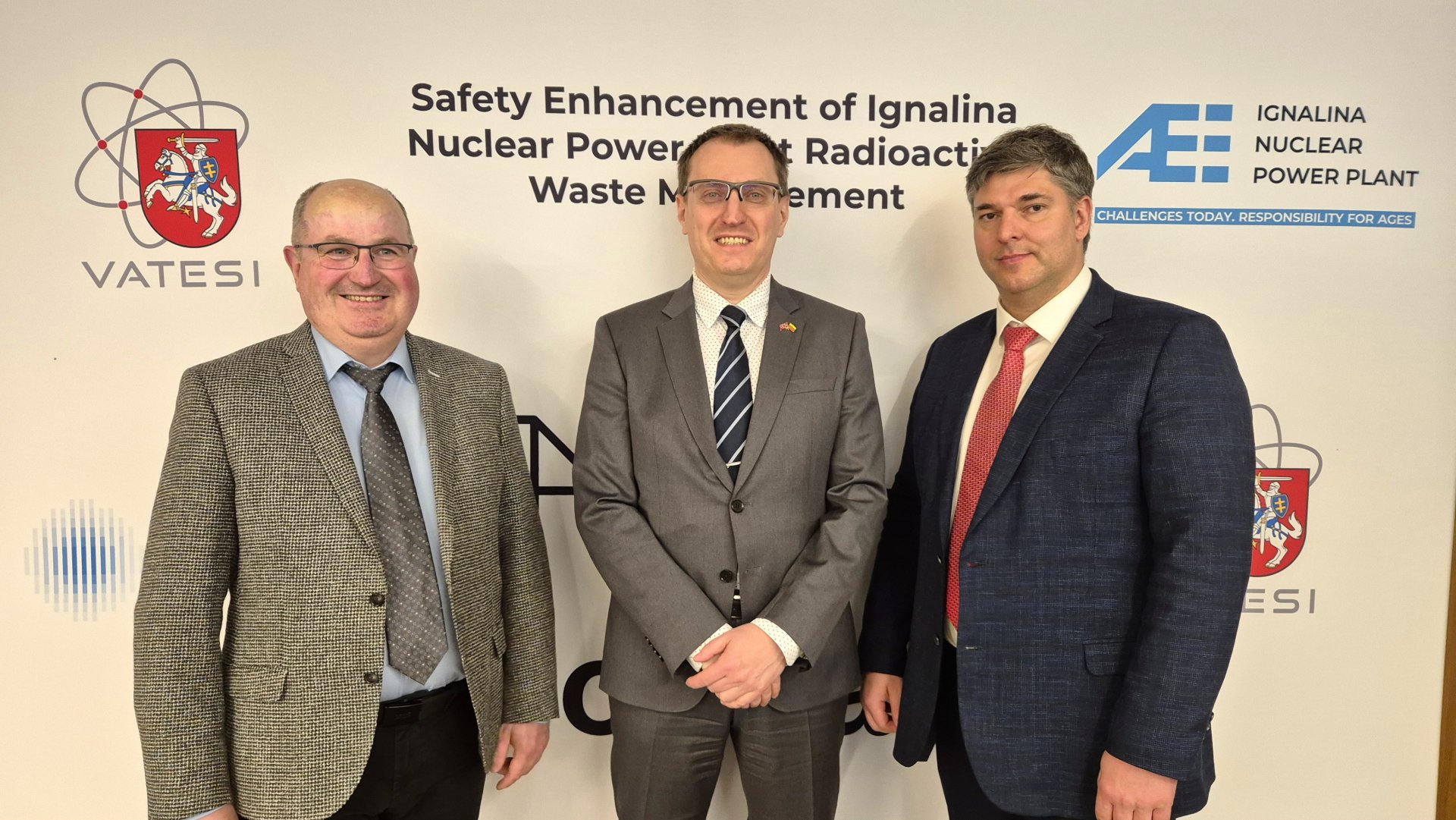Trys vyrai stovi prie informacinės sienelės su Ignalinos atominės elektrinės ir VATESI logotipais, aptariant branduolinių atliekų saugos projektą.