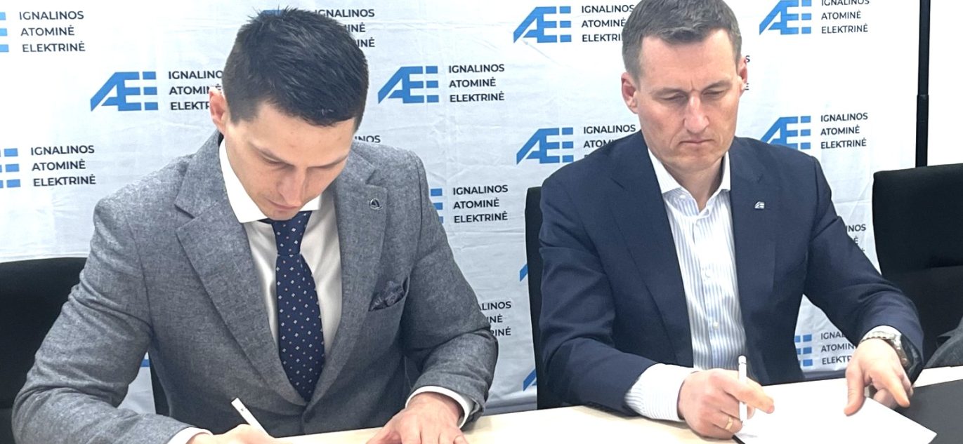 Du vyrai pasirašo sutartį Ignalinos atominės elektrinės fone, dėvi kostiumus, fonas su AE logotipais.