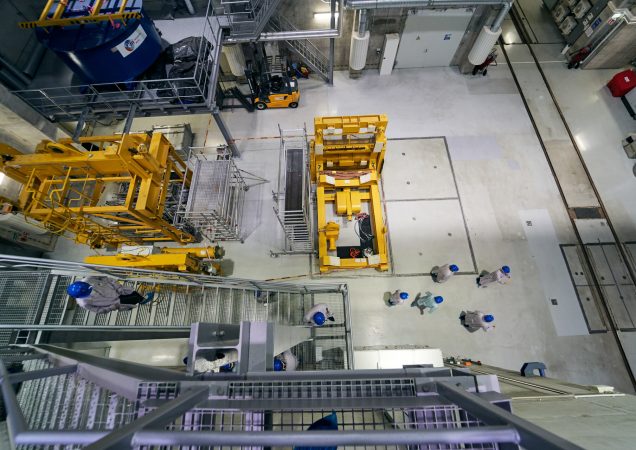 Darbuotojai su šalmais dirba Ignalinos atominės elektrinės viduje prie geltonos įrangos.