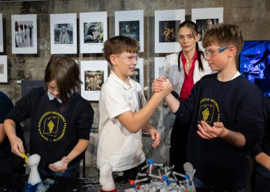 Trys mokiniai atlieka eksperimentus su cheminėmis medžiagomis, du iš jų draugiškai spaudžia rankas, fone – edukacijos vadovė ir nuotraukų paroda.