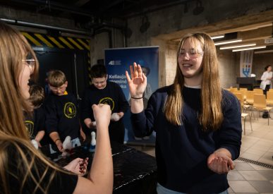 Jaunimas su apsauginiais akiniais dalyvauja edukacinėje veikloje Ignalinos atominėje elektrinėje, prie stalo žaismingai demonstruodami rankų gestus.