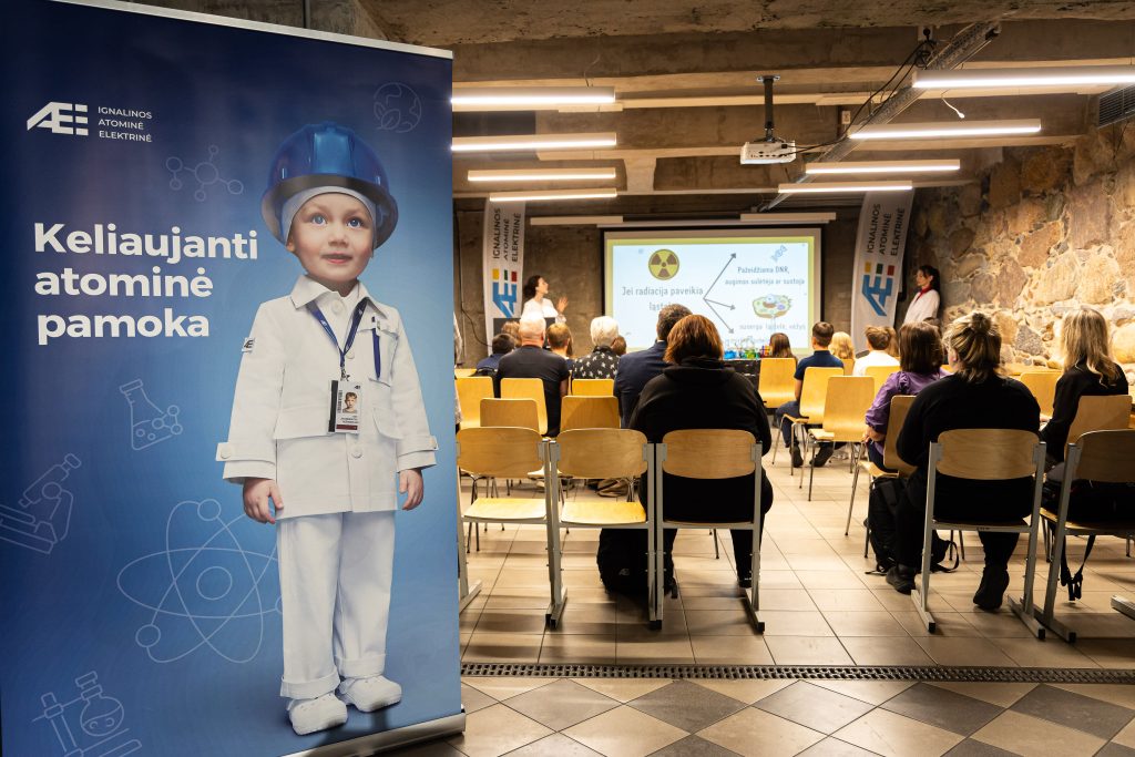Ignalinos atominės elektrinės renginyje vyksta edukacinė pamoka, kurioje dalyviai sėdi priešais ekraną, o šone matomas stendas su užrašu Keliaujanti atominė pamoka.