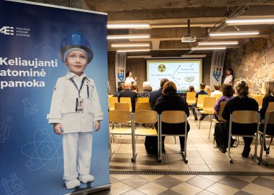 Ignalinos atominės elektrinės renginyje vyksta edukacinė pamoka, kurioje dalyviai sėdi priešais ekraną, o šone matomas stendas su užrašu Keliaujanti atominė pamoka.