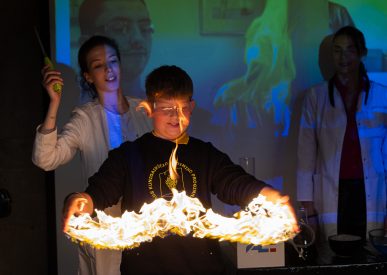 Mokinys su tamsiu mokyklos uniformos megztiniu Ignalinos atominės elektrinės edukaciniame renginyje atlieka bandymą, kai rankų aukštyje sukurta liepsnos ugnies juosta, šalia stebi mokslininkės su baltais chalatais.
