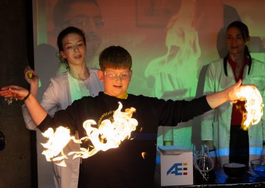 Mokinys su tamsiu mokyklos uniformos megztiniu Ignalinos atominės elektrinės edukaciniame renginyje atlieka bandymą, kai rankų aukštyje sukurta liepsnos ugnies juosta, šalia stebi mokslininkės su baltais chalatais.