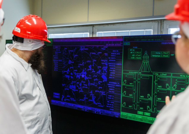 Darbuotojai su raudonais šalmais ir baltais apsauginiais drabužiais stebi valdymo pulto ekranus Ignalinos atominėje elektrinėje.