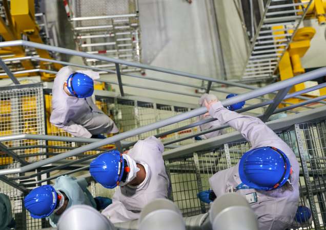 Darbuotojai su mėlynais šalmais ir apsauginiais rūbais leidžiasi metaliniais laiptais pramoninėje Ignalinos atominės elektrinės patalpoje.