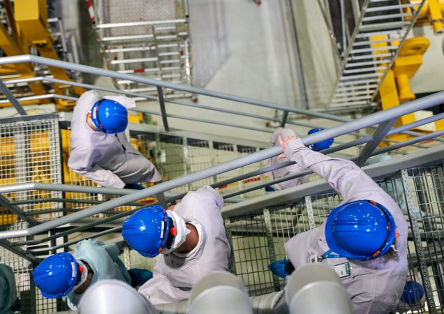Darbuotojai su mėlynais šalmais ir apsauginiais drabužiais eina metalinėmis laiptais jėgainės patalpoje.