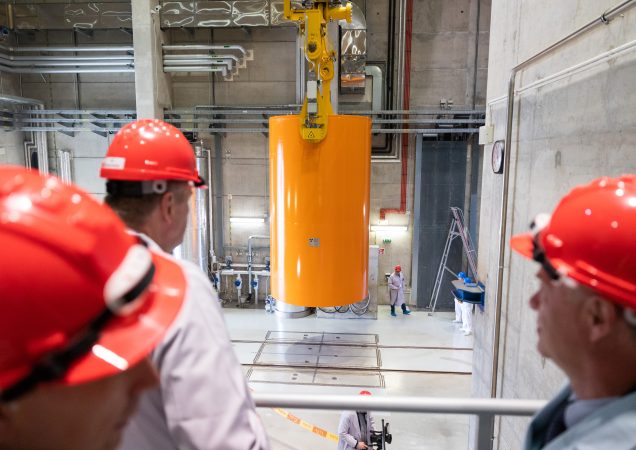 Darbuotojai su raudonais šalmais stebi, kaip kėlimo įranga Ignalinos atominėje elektrinėje pakelia didelę oranžinę radioaktyviųjų atliekų saugojimo talpą.