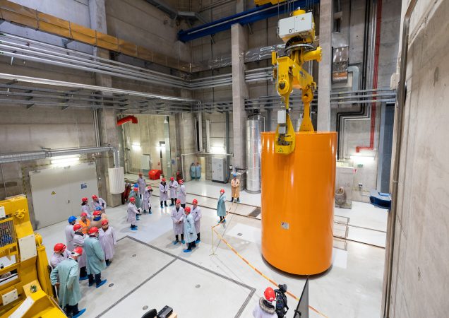 Darbuotojai su šalmais ir apsauginiais drabužiais Ignalinos atominėje elektrinėje stebi, kaip kėlimo kranas laiko didelę oranžinę radioaktyviųjų atliekų saugojimo talpą.