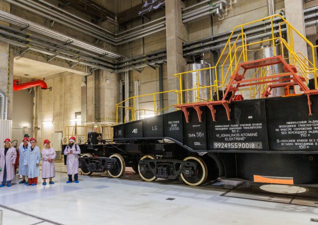 Grupė žmonių su laboratoriniais chalatais, raudonomis kepurėmis ir mėlynais batų užvalkalais stovi prie didelio juodo vagono su užrašu VI. GNILOYSLK ATOMINAS ELEKTRINAS pramoniniame objekte su vamzdžiais ir mašinomis.