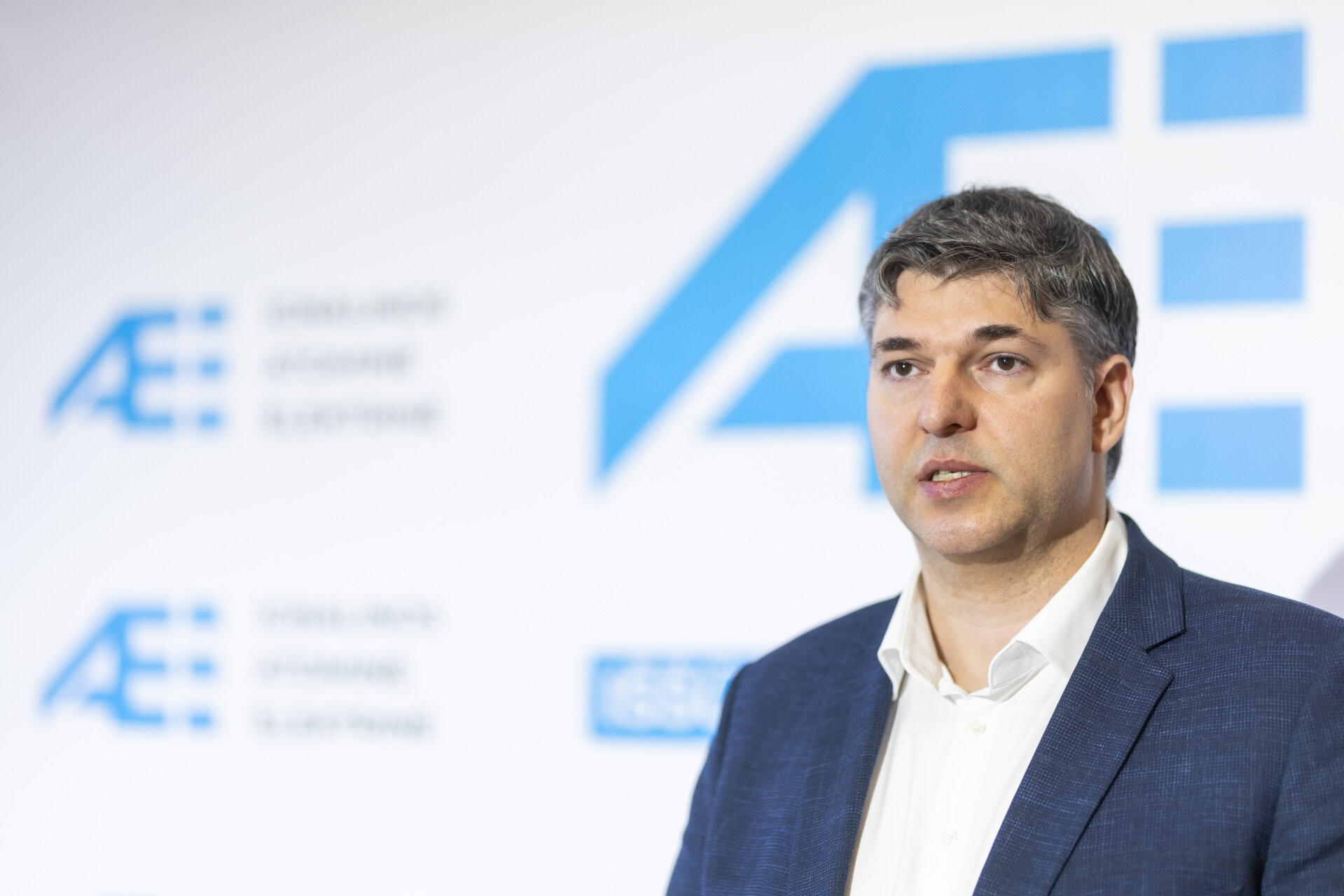 Vyras tamsiu kostiumu stovi prieš AE logotipą pristatymo metu.