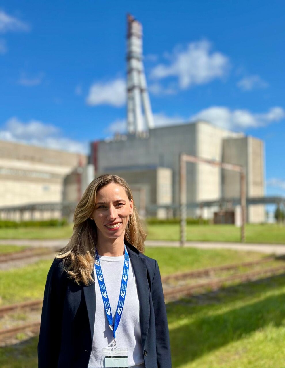 Rasa Danielievičienė stovi lauke prie Ignalinos atominės elektrinės pastato su matomu reaktoriaus kaminu fone.