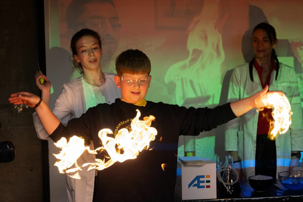 Mokinys su tamsiu mokyklos uniformos megztiniu Ignalinos atominės elektrinės edukaciniame renginyje atlieka bandymą, kai rankų aukštyje sukurta liepsnos ugnies juosta, šalia stebi mokslininkės su baltais chalatais.
