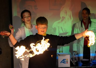 Mokinys su tamsiu mokyklos uniformos megztiniu Ignalinos atominės elektrinės edukaciniame renginyje atlieka bandymą, kai rankų aukštyje sukurta liepsnos ugnies juosta, šalia stebi mokslininkės su baltais chalatais.
