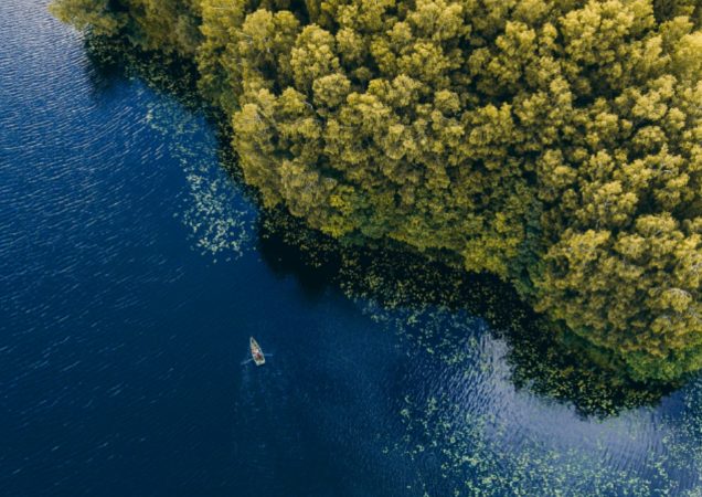 Orto nuotrauka: žalias miško masyvas šalia ežero, kuriame matomas mažas laivas plaukiantis mėlynu vandeniu.