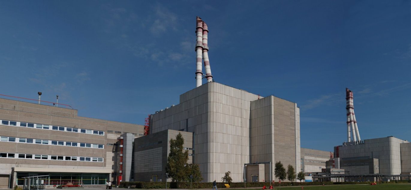 Ignalinos atominės elektrinės pastatų kompleksas su aukštais raudonai baltais dūmų išmetimo kaminais giedro dangaus fone.