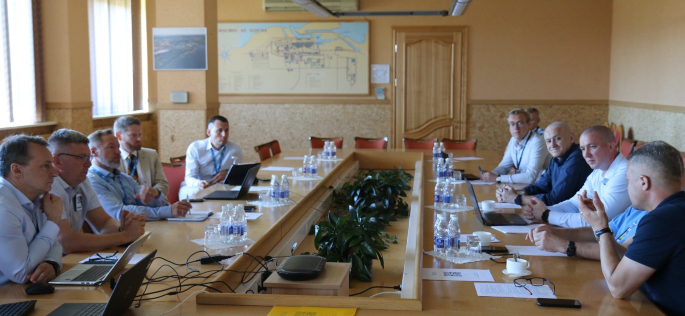 Ignalinos atominės elektrinės darbuotojų pasitarimas konferencijų salėje, dalyviai sėdi prie ilgo stalo su nešiojamaisiais kompiuteriais ir dokumentais.