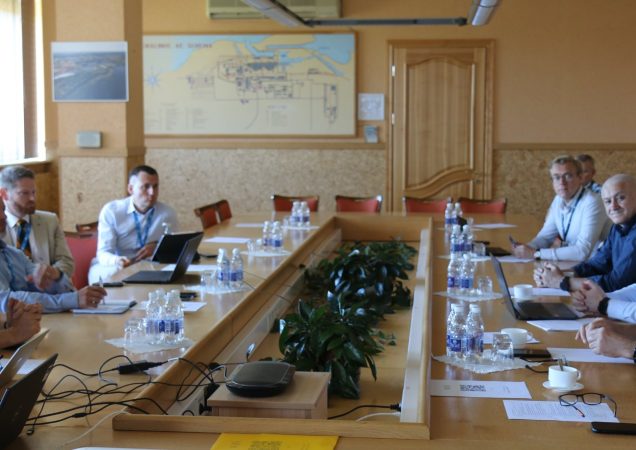 Ignalinos atominės elektrinės darbuotojų pasitarimas konferencijų salėje, dalyviai sėdi prie ilgo stalo su nešiojamaisiais kompiuteriais ir dokumentais.
