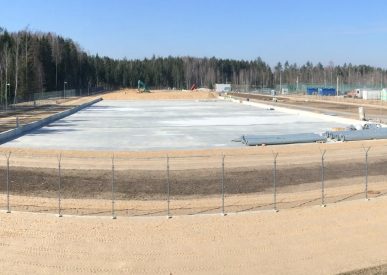 Betoninė aikštelė uždarojoje statybvietėje, aptvertoje metaline tvora su spygliuota viela, fone matosi miškas, kranai ir pagalbiniai pastatai.