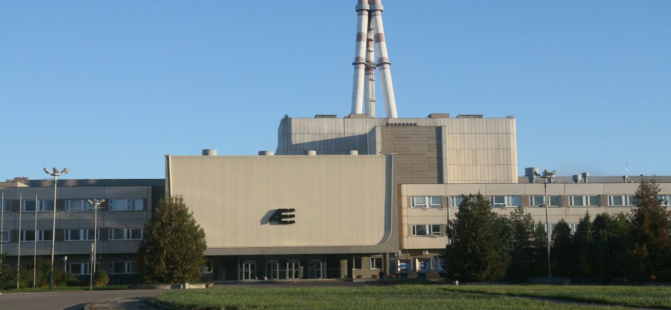 Ignalinos atominė elektrinė su dviem reaktoriaus bokštais, matoma saulėtą dieną iš priekio. Pastato fasade – AE logotipas.