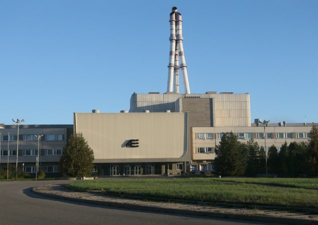 Ignalinos atominė elektrinė su dviem reaktoriaus bokštais, matoma saulėtą dieną iš priekio. Pastato fasade – AE logotipas.