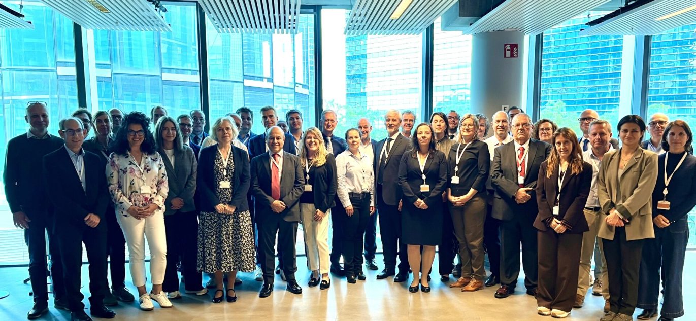 Grupė žmonių stovi kartu konferencijos salėje Briuselyje prie didelių langų.