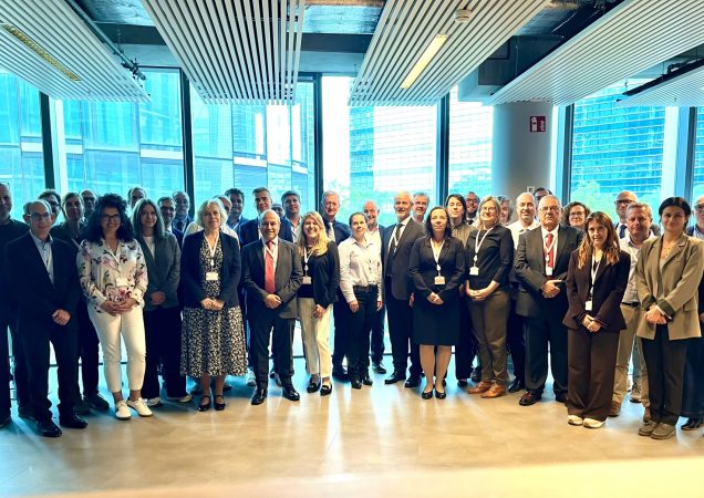 Grupė žmonių stovi kartu konferencijos salėje Briuselyje prie didelių langų.