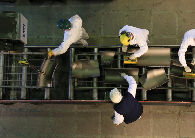 Darbuotojai su apsauginiais šalmais dirba prie metalo detalių konvejerio linijos