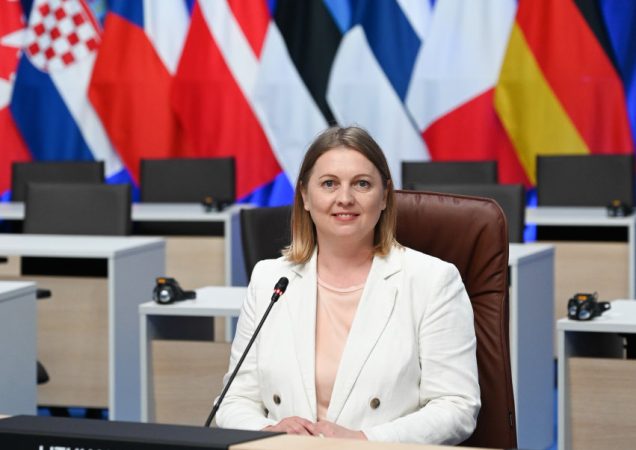 Moteris baltais drabužiais sėdi prie mikrofono tarptautinėje konferencijoje, fone – įvairių valstybių vėliavos, tarp jų Lietuvos, Vokietijos, Prancūzijos ir kitų. Ant stalo lentelė su užrašu „Lithuania“.