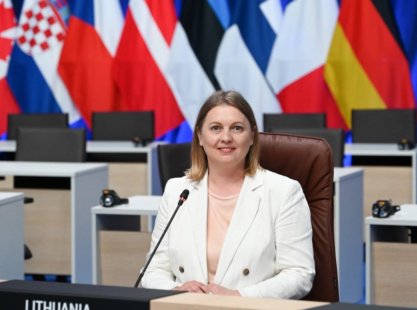 Moteris baltais drabužiais sėdi prie mikrofono tarptautinėje konferencijoje, fone – įvairių valstybių vėliavos, tarp jų Lietuvos, Vokietijos, Prancūzijos ir kitų. Ant stalo lentelė su užrašu „Lithuania“.