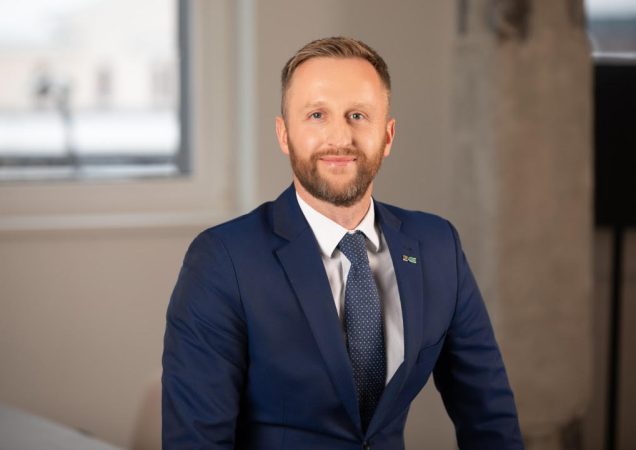 Komiteto narys vilkintis tamsiai mėlyną kostiumą ir taškuotą kaklaraištį sėdi prie stalo, fone matosi langas.