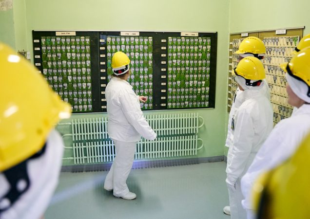 Keli žmonės baltais apsauginiais kostiumais ir geltonais šalmais stovi kambaryje ir stebi lentą, padengtą eilėmis žalių žymeklių arba kortelių. Vienas asmuo rodo į lentą ir tarsi kažką aiškina.