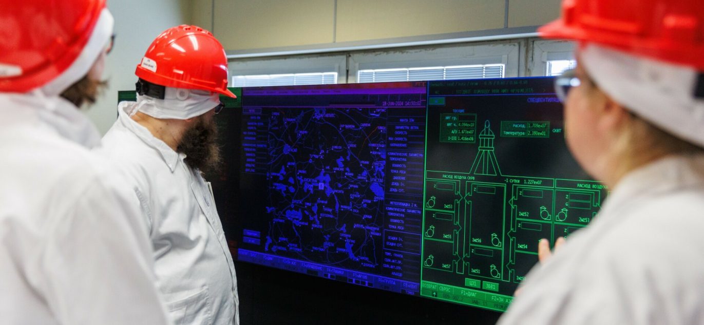 Darbuotojai su raudonais šalmais ir baltais apsauginiais drabužiais stebi valdymo pulto ekranus Ignalinos atominėje elektrinėje.