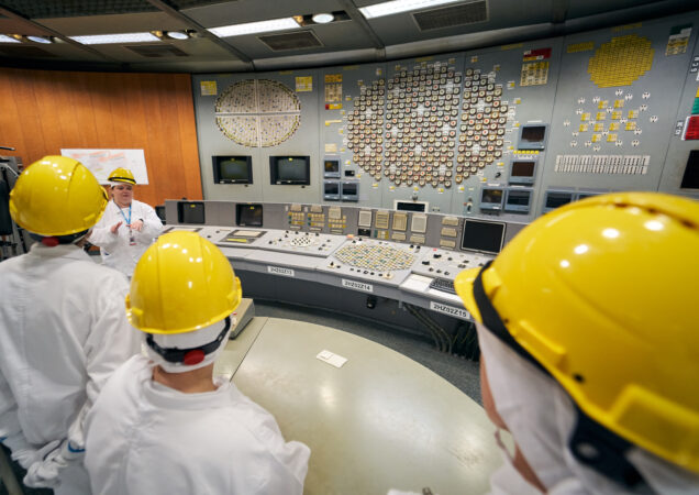 Žmonės su geltonais šalmais ir apsauginiais kostiumais klausosi instruktažo elektrinės viduje