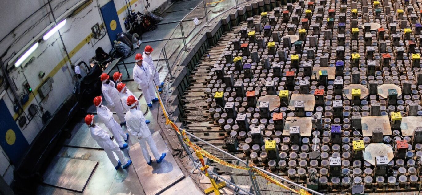 Reaktoriaus salėje matomas Ignalinos atominės elektrinės reaktorius, šalia jo grupė baltai specialiais kostiumais apsirengusių žmonių su raudonais šalmais