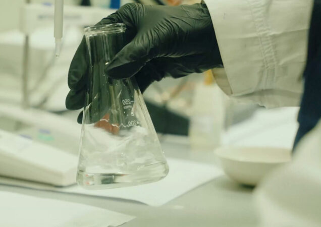 Laboratorijoje atliekamas tikslus cheminis tyrimas – ranka su apsaugine pirštine laiko kolbą su skaidriu skysčiu